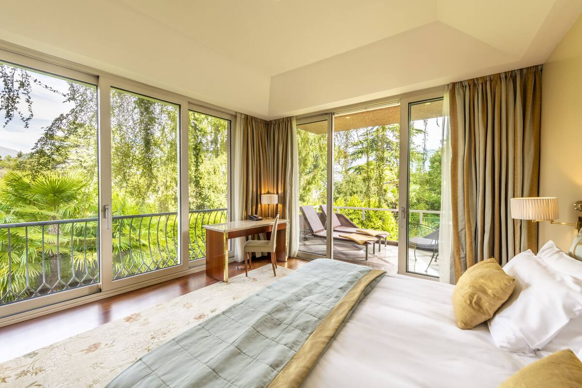 A spacious bedroom with a large bed, a desk, and a balcony with a view of lush greenery.