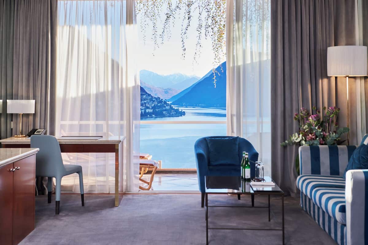 A hotel room with a desk, two chairs, a sofa, and a view of mountains and a lake.