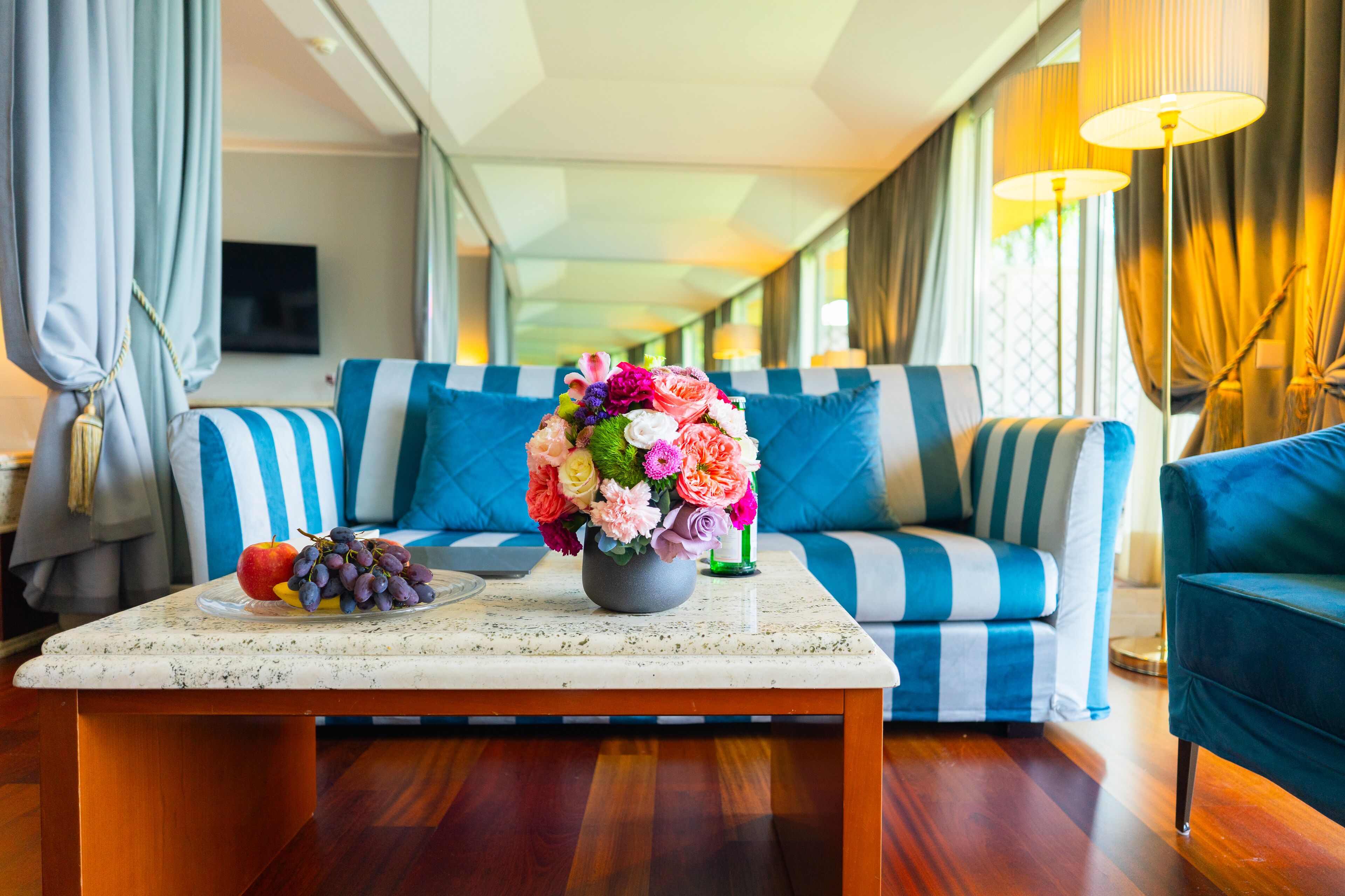 A modern living room with a striped sofa, a coffee table with fruits and flowers, and a TV in the background.