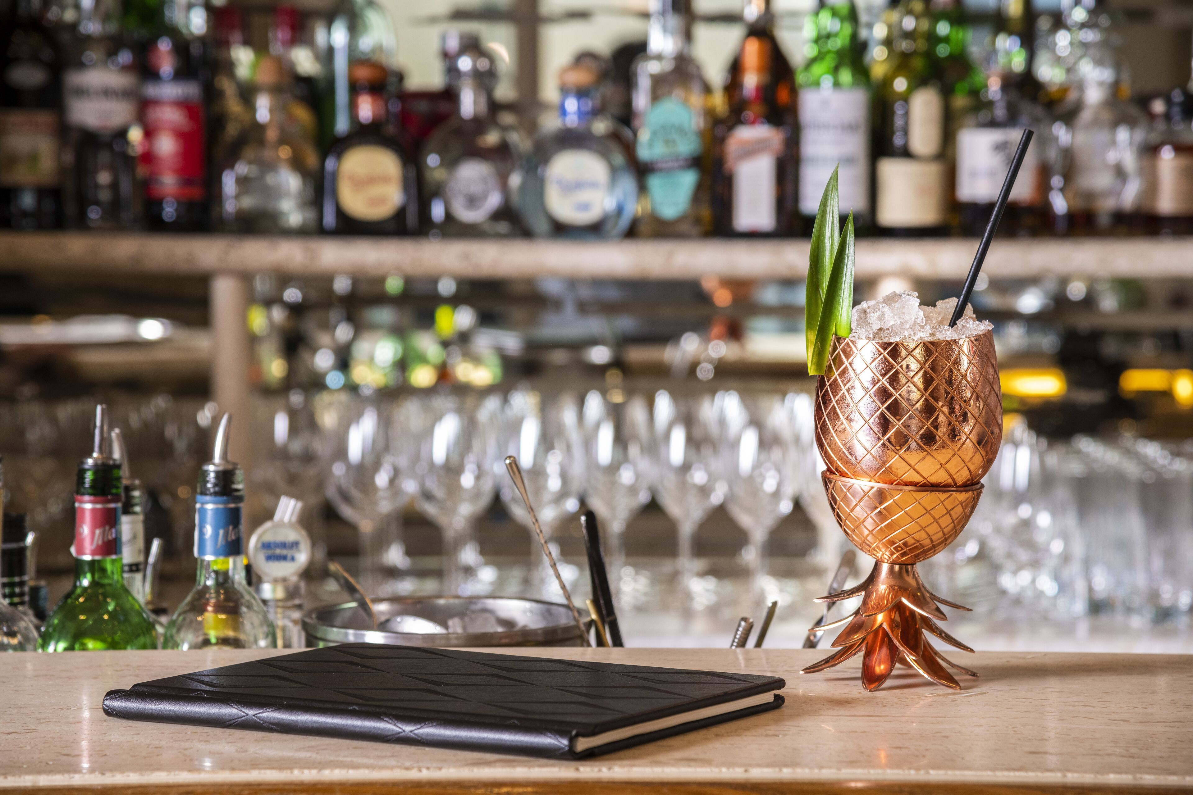 Una zona de bar con un vaso de cóctel de cobre sobre una bandeja, un cuaderno y varias botellas de alcohol.