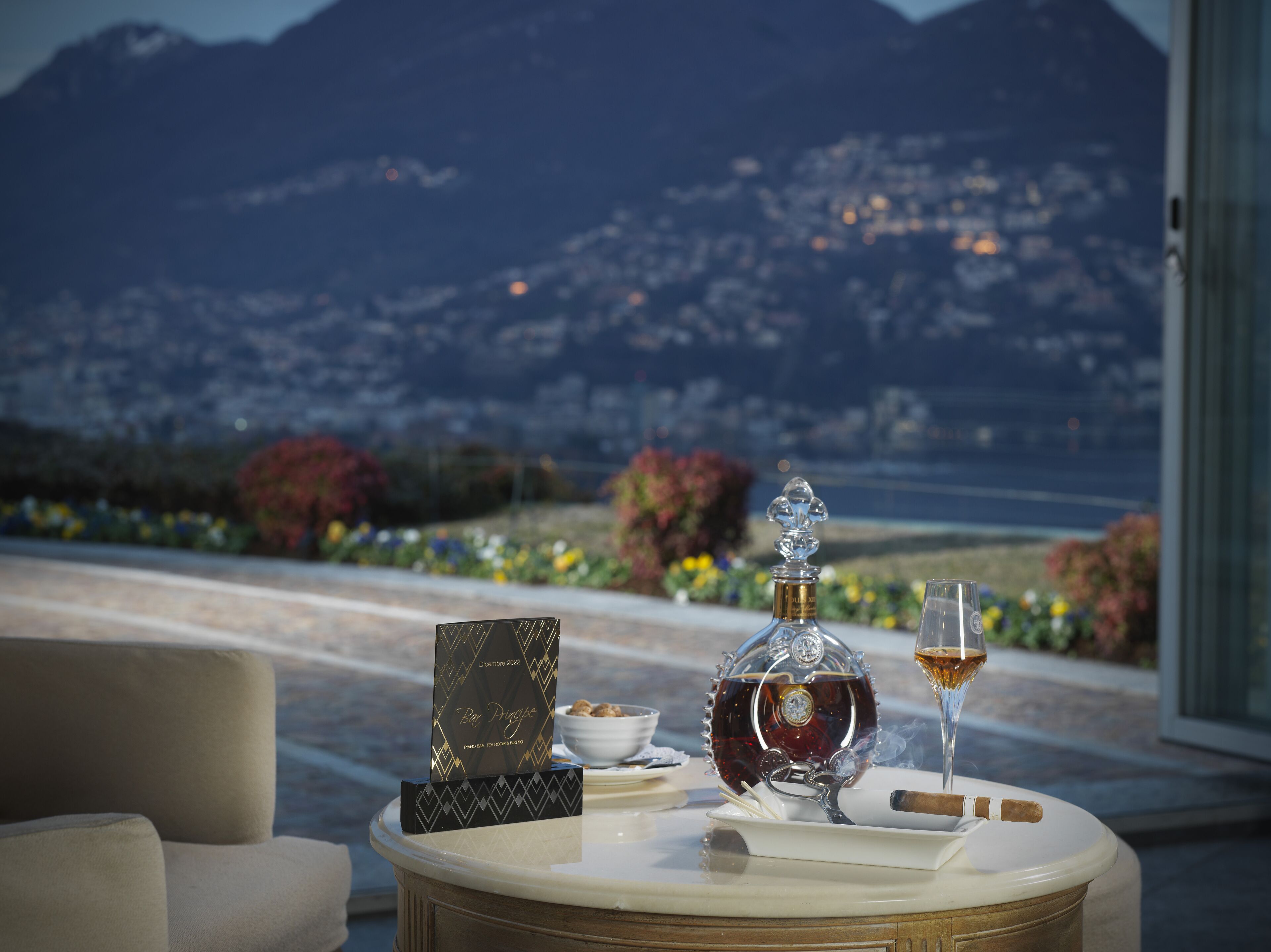 Ein Tisch mit einer Spirituosenflasche, einem Weinglas, einer Zigarre und einer Speisekarte auf einer Terrasse mit Bergblick.