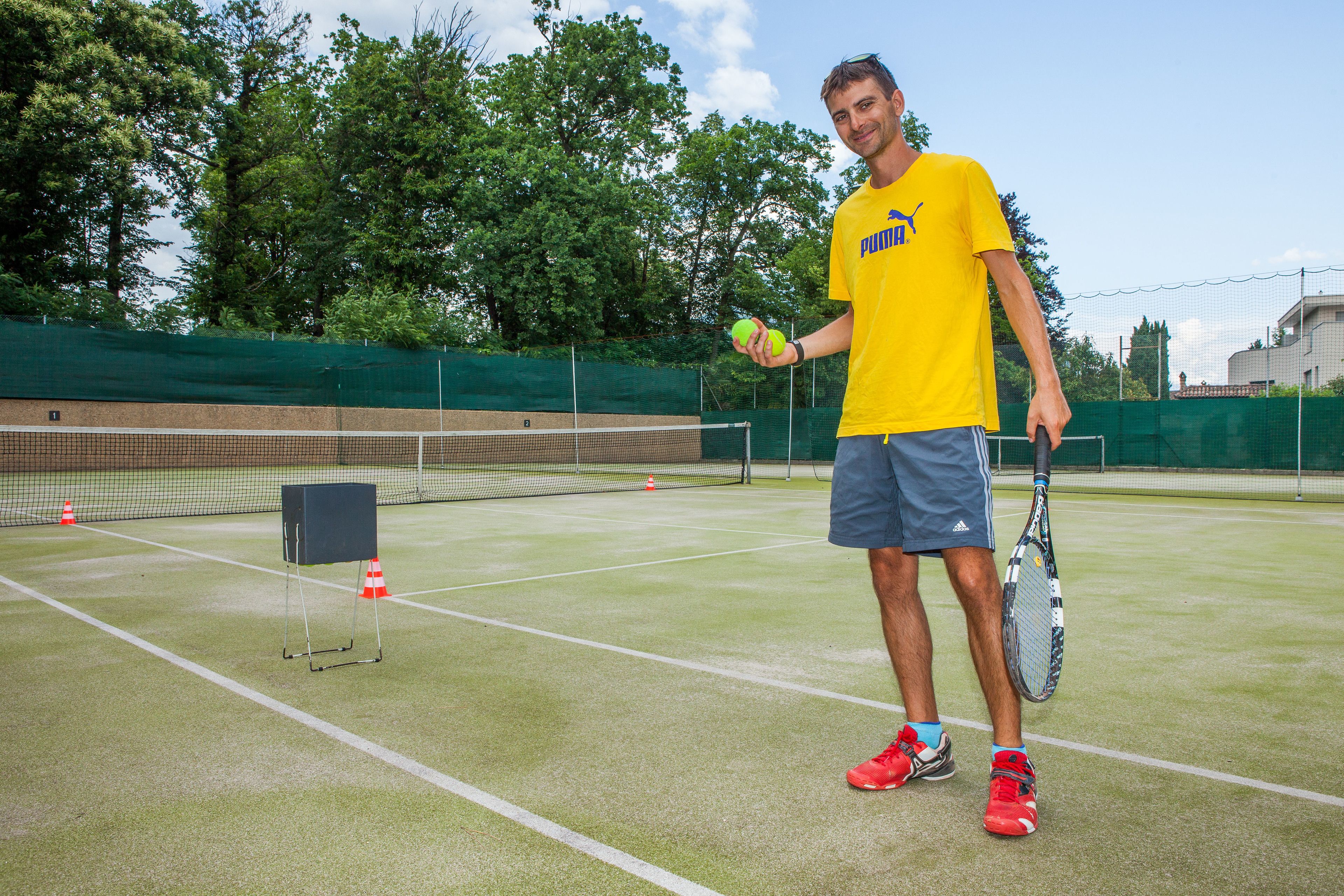 A tennis court with a net, a player holding a tennis racket and balls, and a green fence in the background.