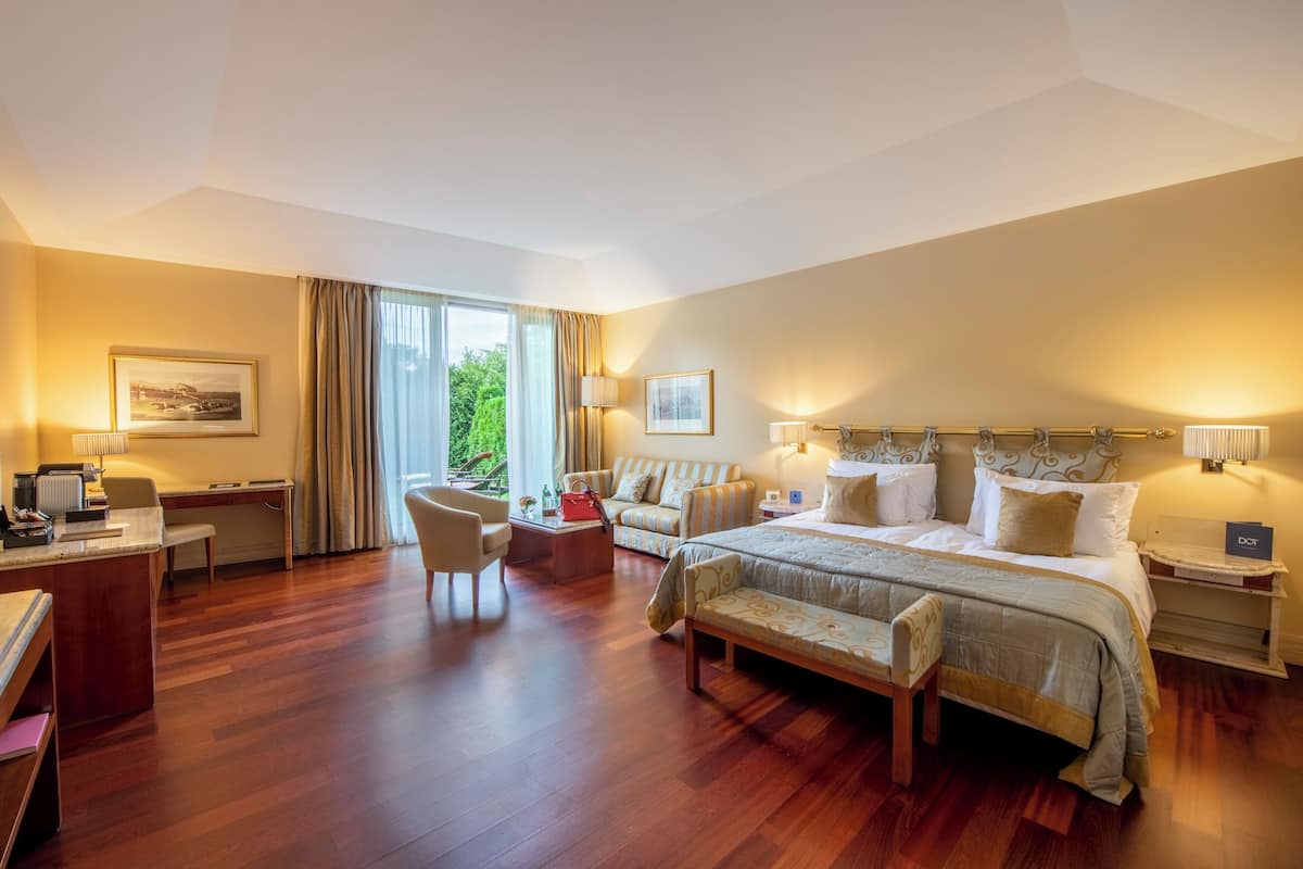 A spacious hotel room with a large bed, a desk, a chair, a sofa, and a view of greenery outside.
