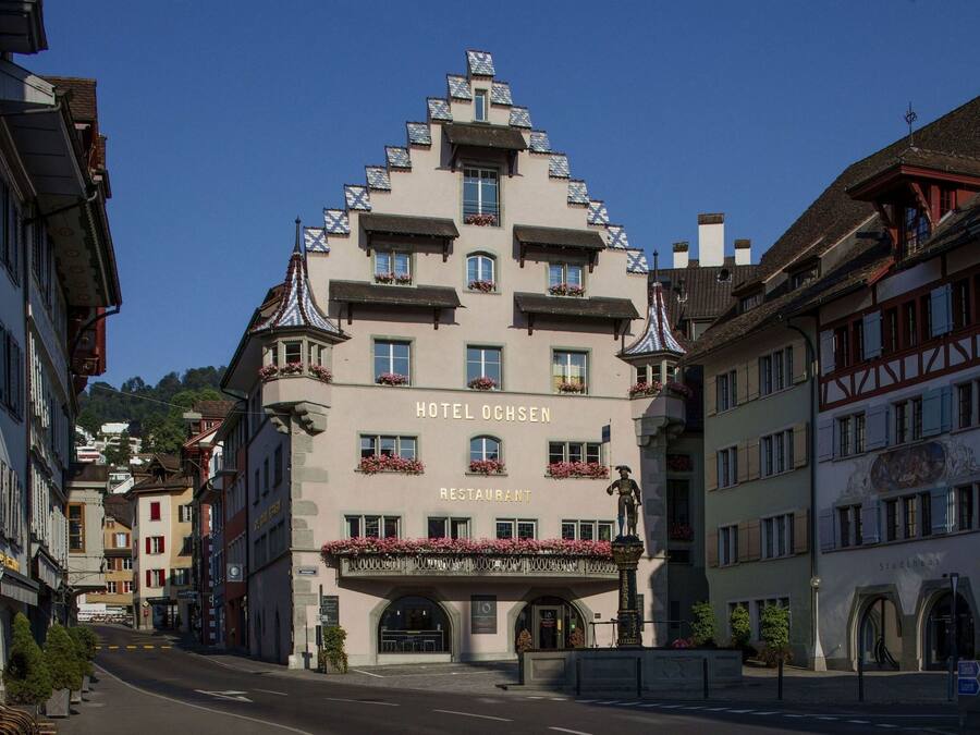 Un hôtel reconnaissable à son enseigne bien visible indiquant « HÔTEL OCHSEN RESTAURANT », et qui présente un style architectural traditionnel avec de nombreux balcons et un toit à forte pente.