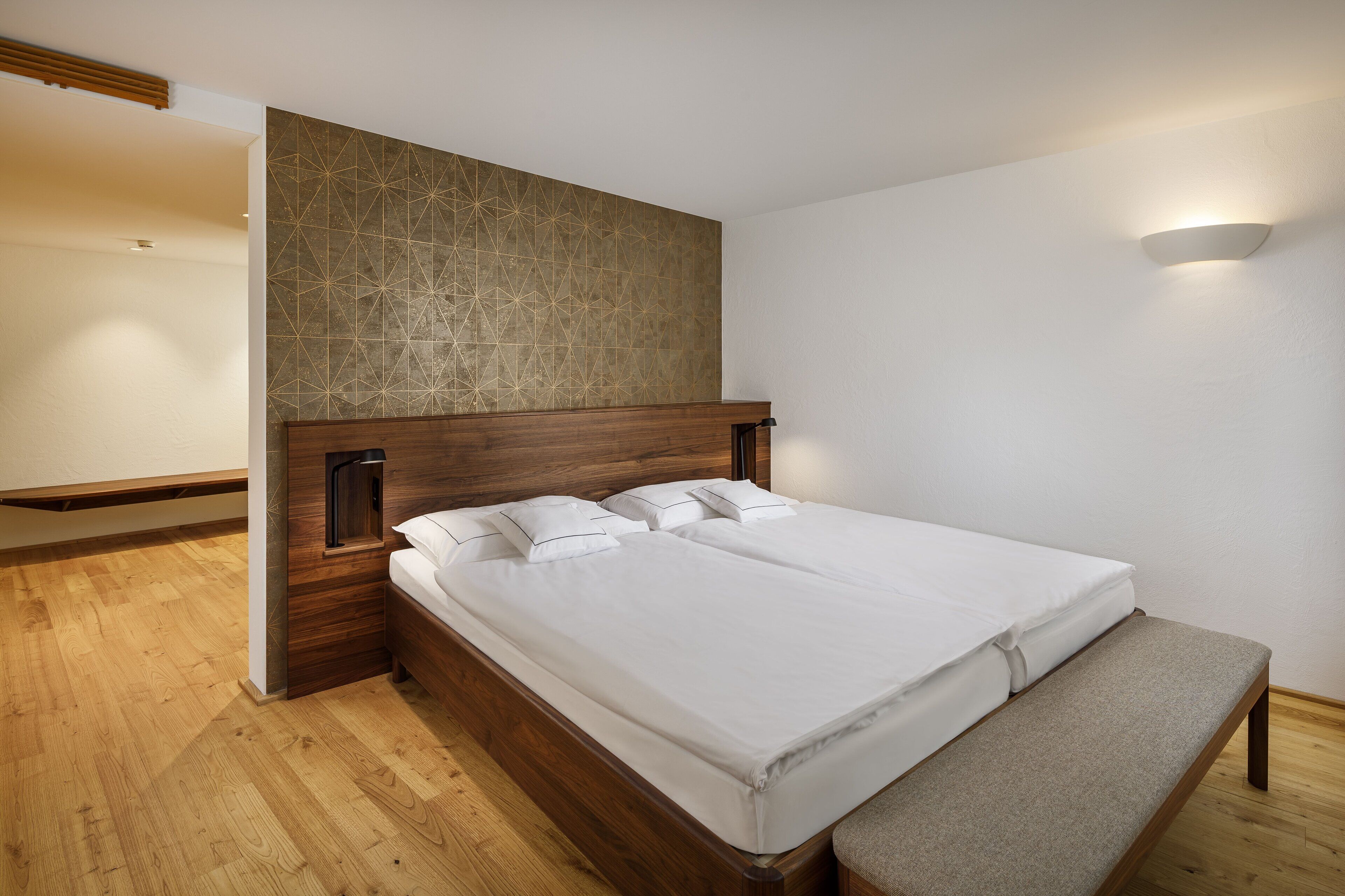 A modern bedroom with a wooden headboard, a bench, and a geometric-patterned wall.