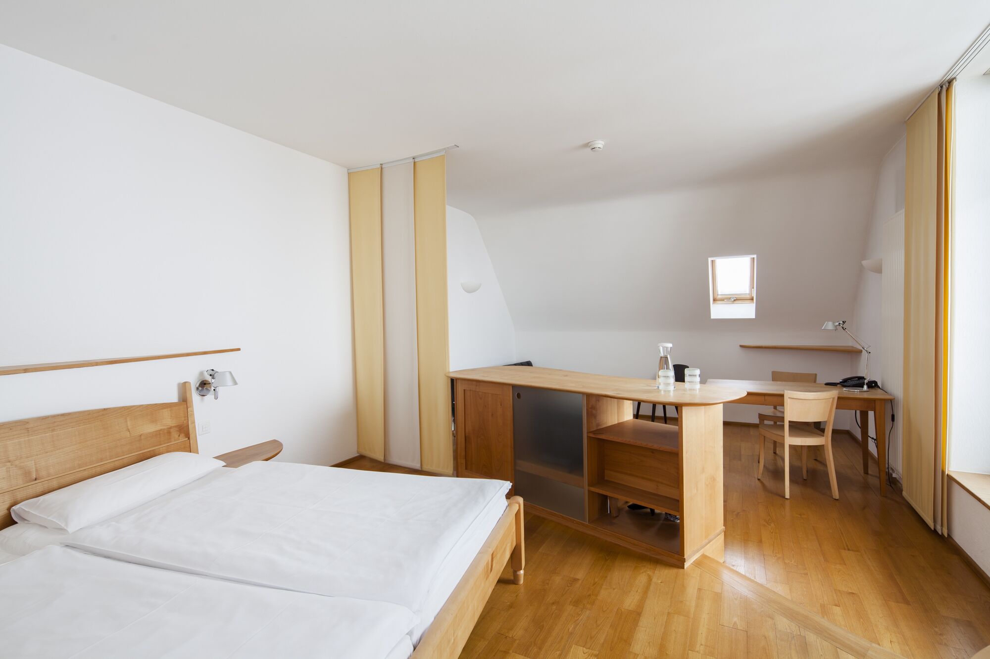 A hotel room with a bed, a desk, and a chair. There is a window providing natural light.