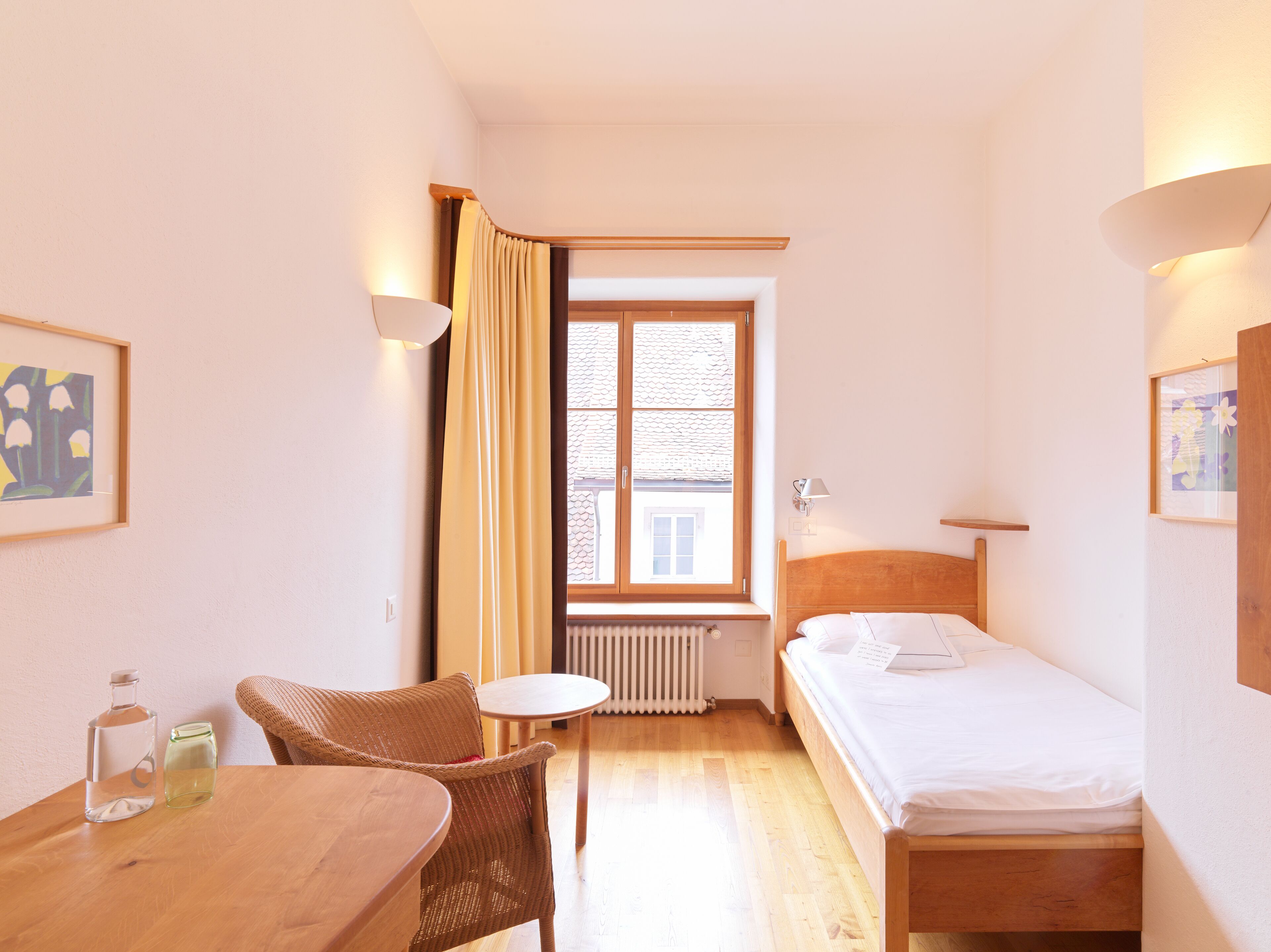 A small, well-lit room with a single bed, a wooden table, wicker chairs, and a window with curtains.