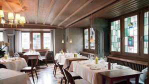 En restaurant med flere spiseborde, store vinduer med farvet glas og trælofter.