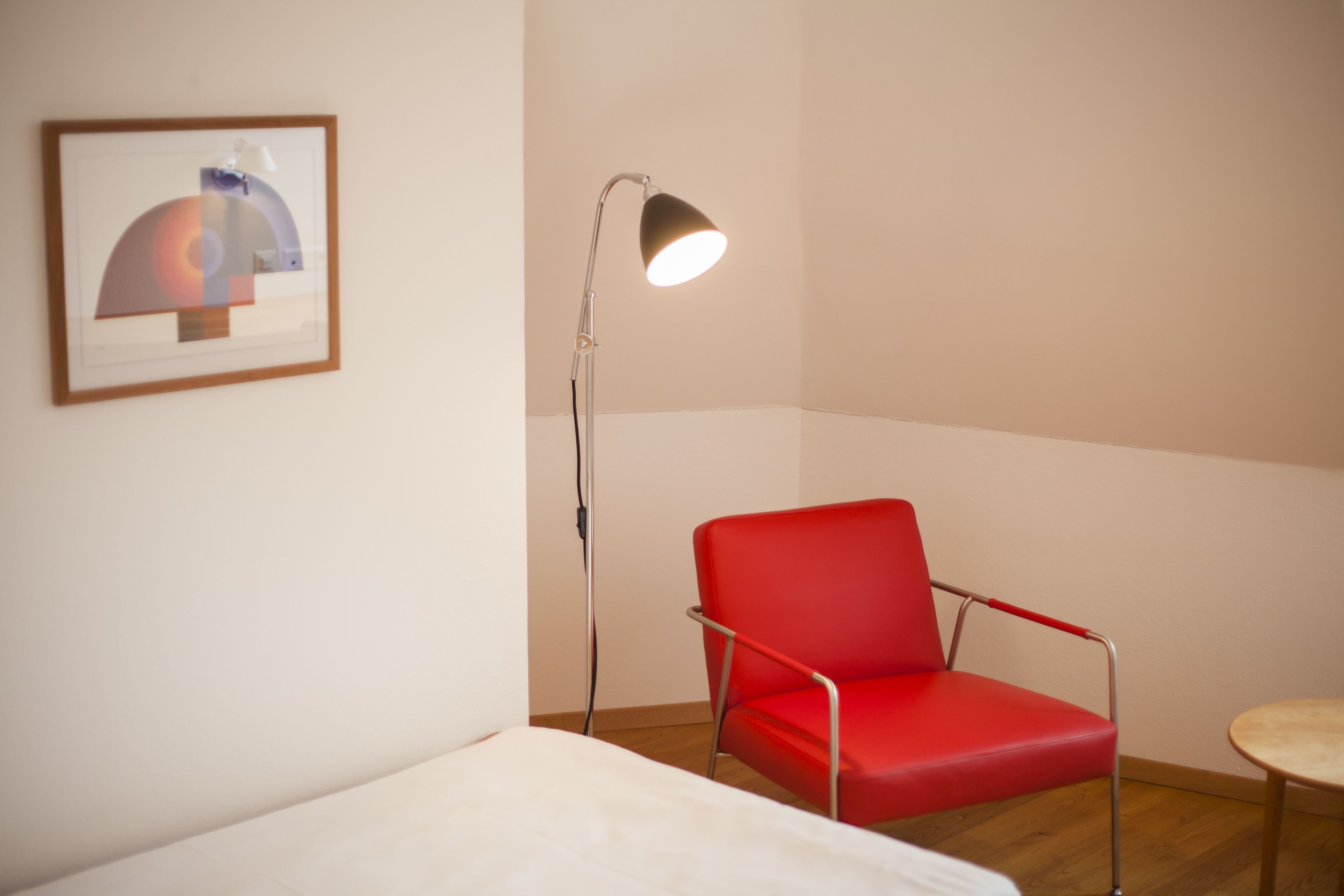 A room with a red chair, a small round table, a floor lamp, and a framed artwork on the wall.