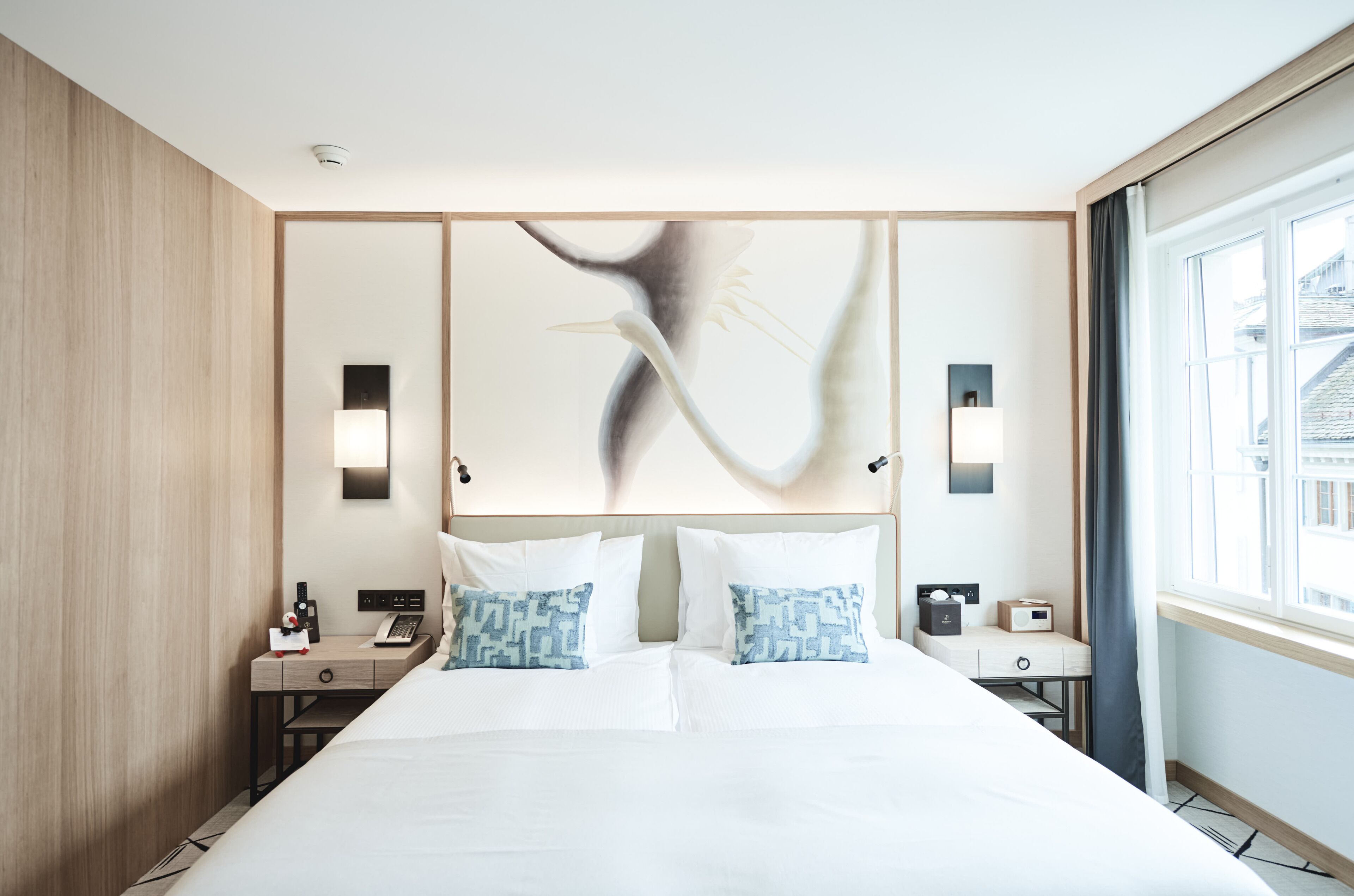 A modern hotel room with a large bed, bedside tables, a desk, and a view of the city through the window.