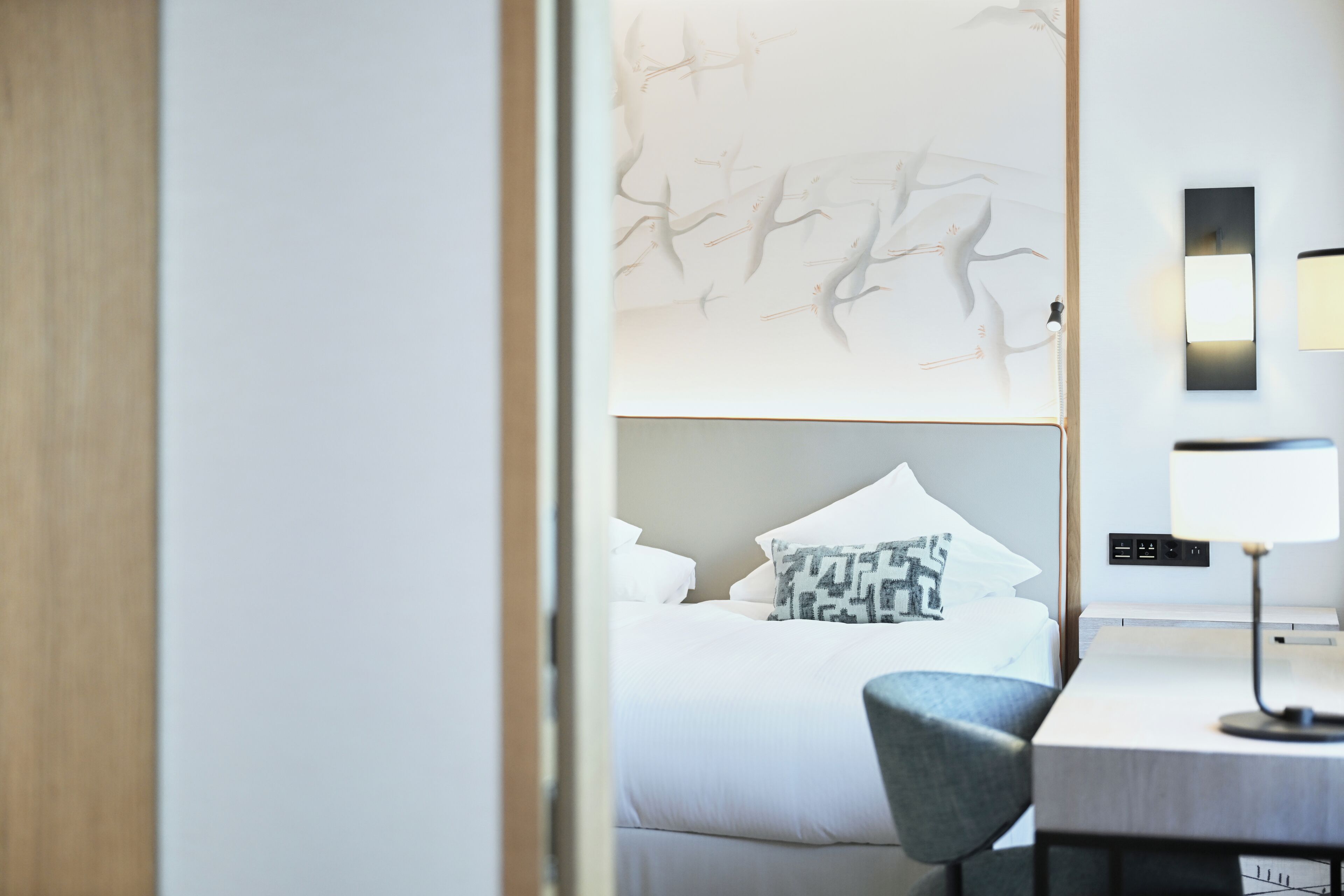 A hotel room with a bed, a desk, a chair, and a wall-mounted lamp.