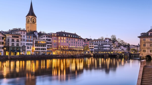 A historic town with a prominent clock tower, reflecting buildings, and a calm river.