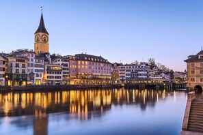 Un pueblo histórico con una imponente torre del reloj, edificios reflejados y un río tranquilo.