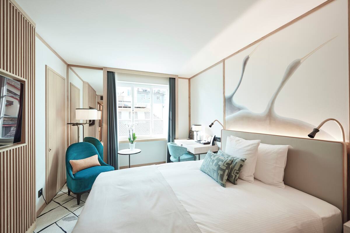 A modern hotel room with a large bed, a desk, a chair, and a view of the city through the window.