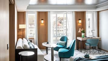 A modern hotel room with a sofa, two armchairs, a small table, a desk, and a view of the city through large windows.