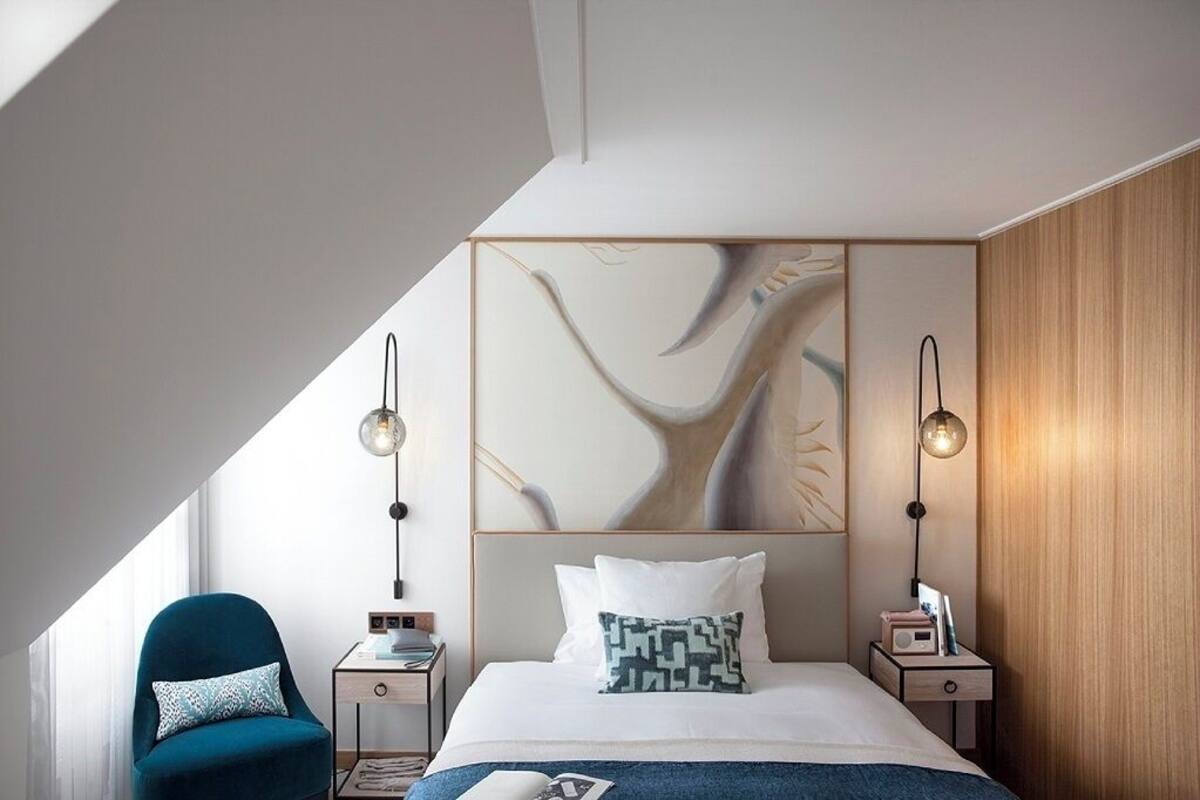 A modern bedroom with a sloped ceiling, a large bed with a geometric headboard, two bedside tables with lamps, a blue armchair, and a wooden accent wall.