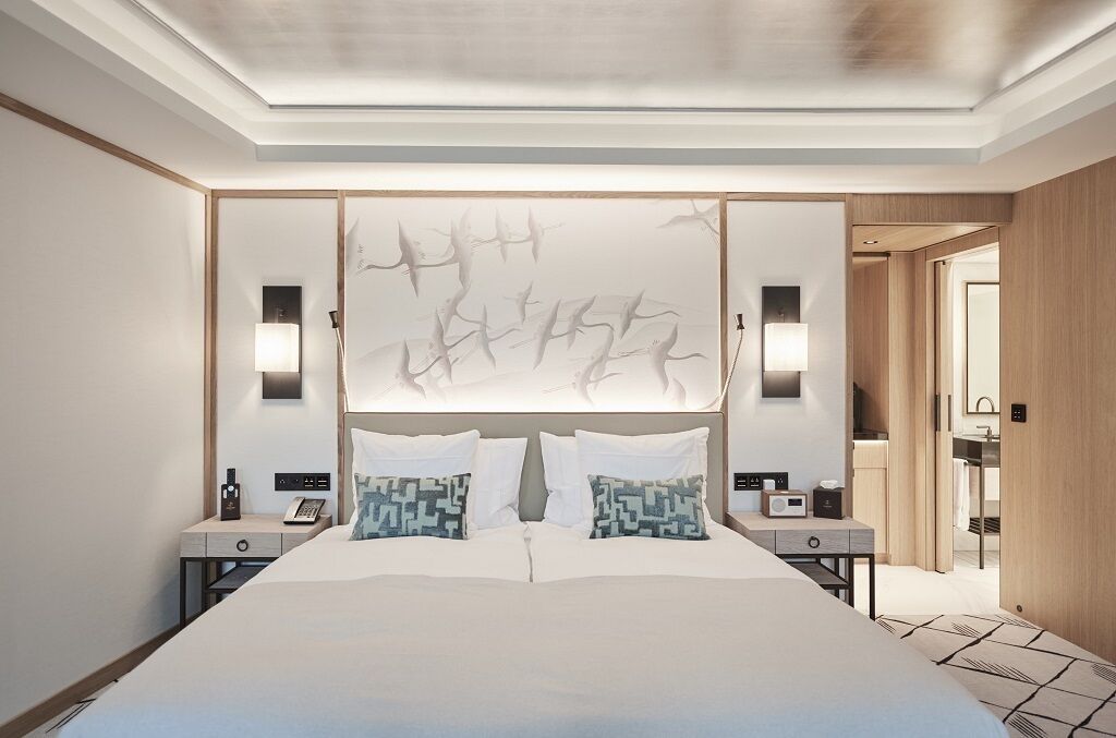 A modern hotel room with a large bed, two bedside tables, a wall-mounted TV, and a bathroom visible through an open door.