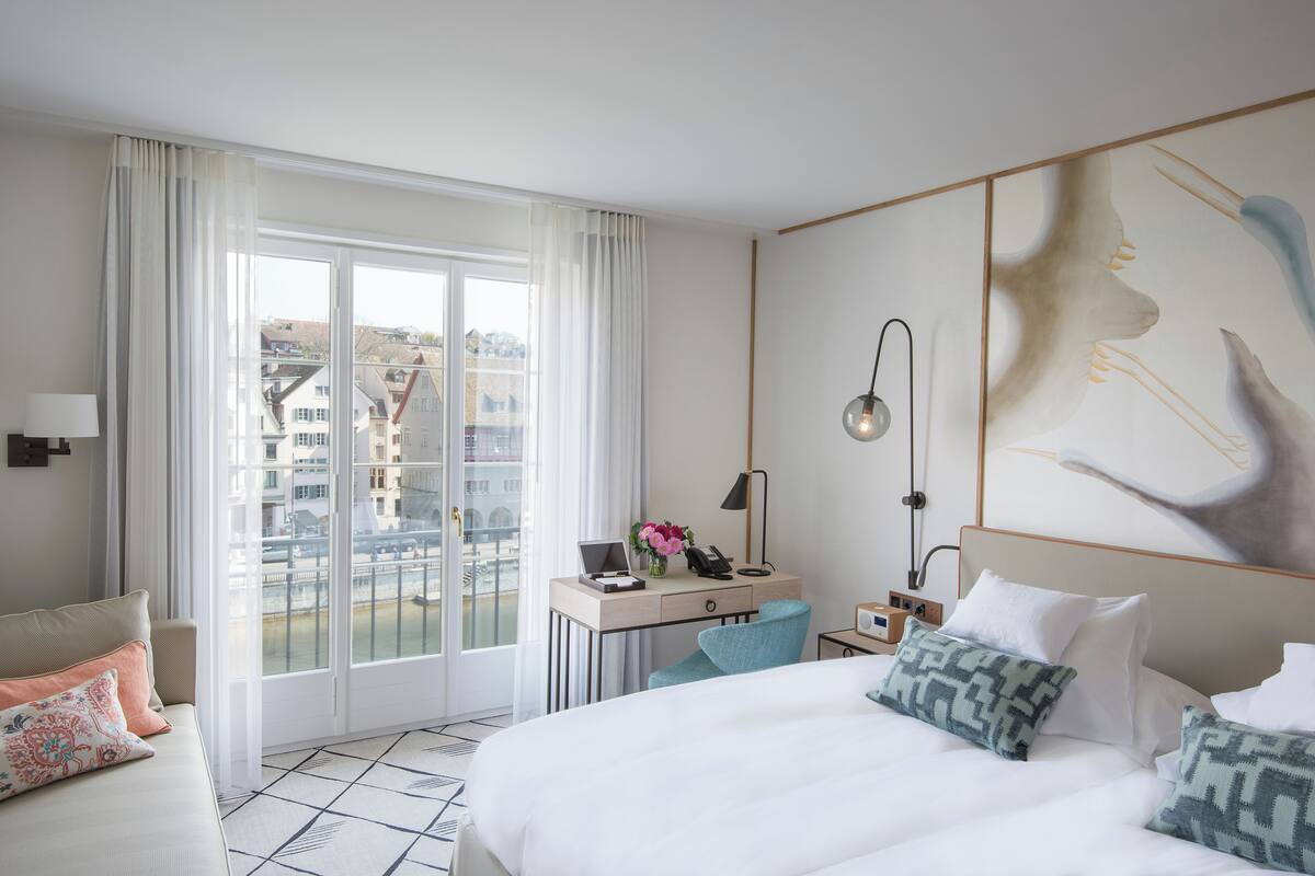 A hotel room with a bed, a desk with a laptop, a chair, and a large window with curtains.