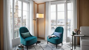 A modern hotel room with large windows, two teal armchairs, a small table with a lamp, and a view of a cityscape.