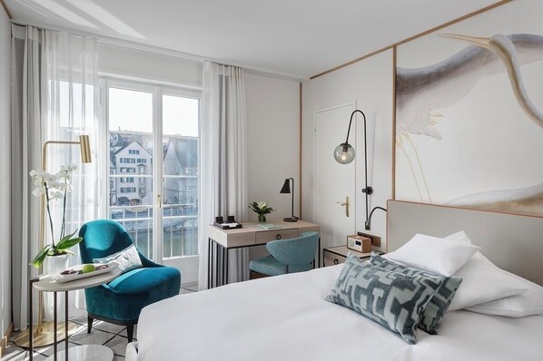 A modern hotel room with a large bed, a desk, a chair, and a view of buildings through the window.
