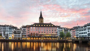 A hotel with a prominent clock tower, a waterfront view, and a reflection in the water.