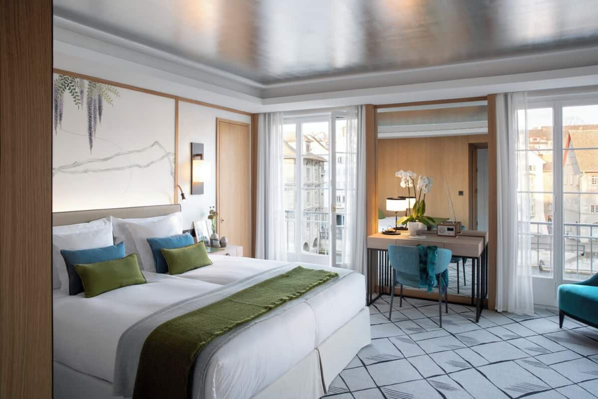 A hotel room with a large bed, a desk, and a view of the city through large windows.