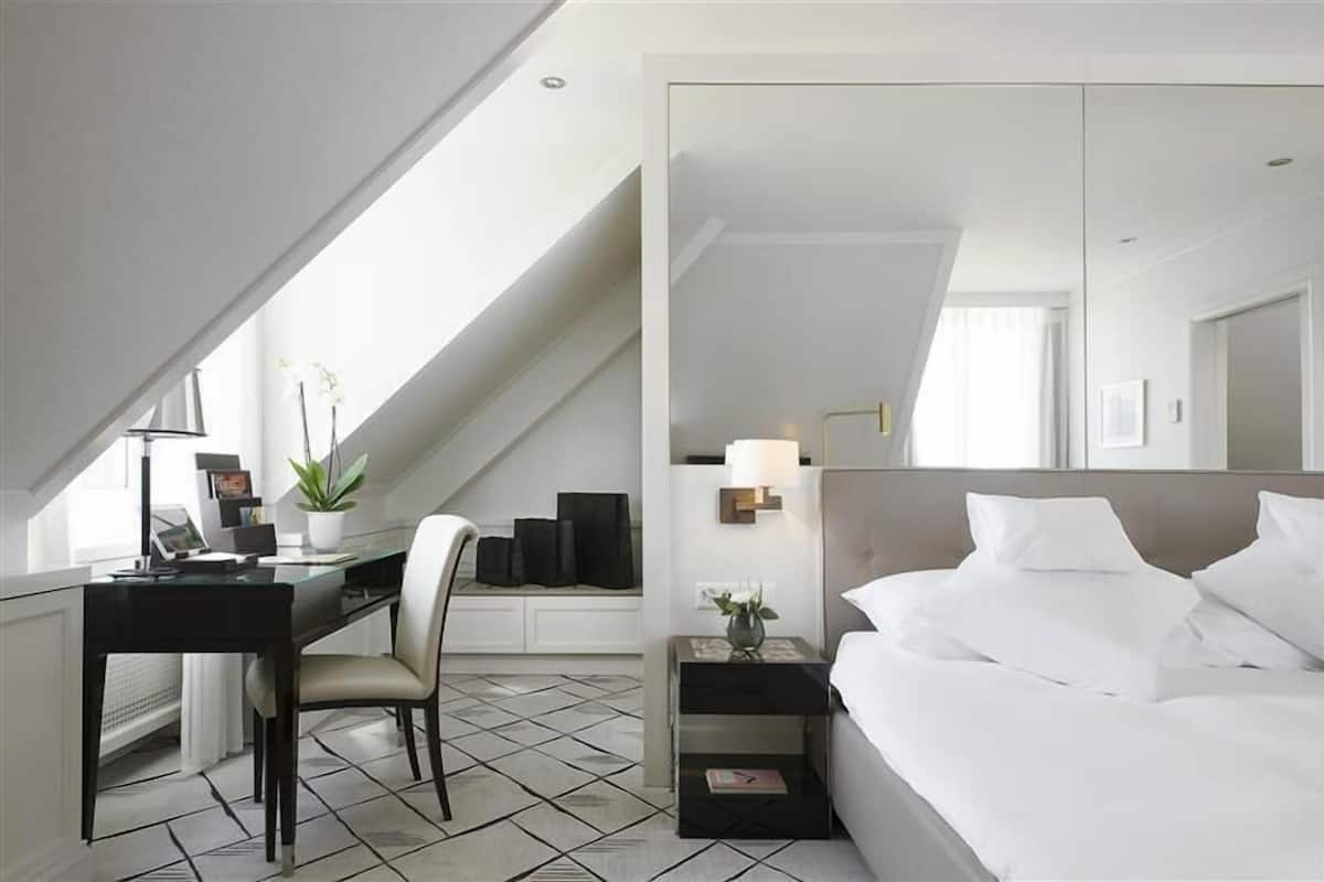 A modern hotel room with a desk, chair, and bed. There is a large mirror above the bed and a small table with a lamp.