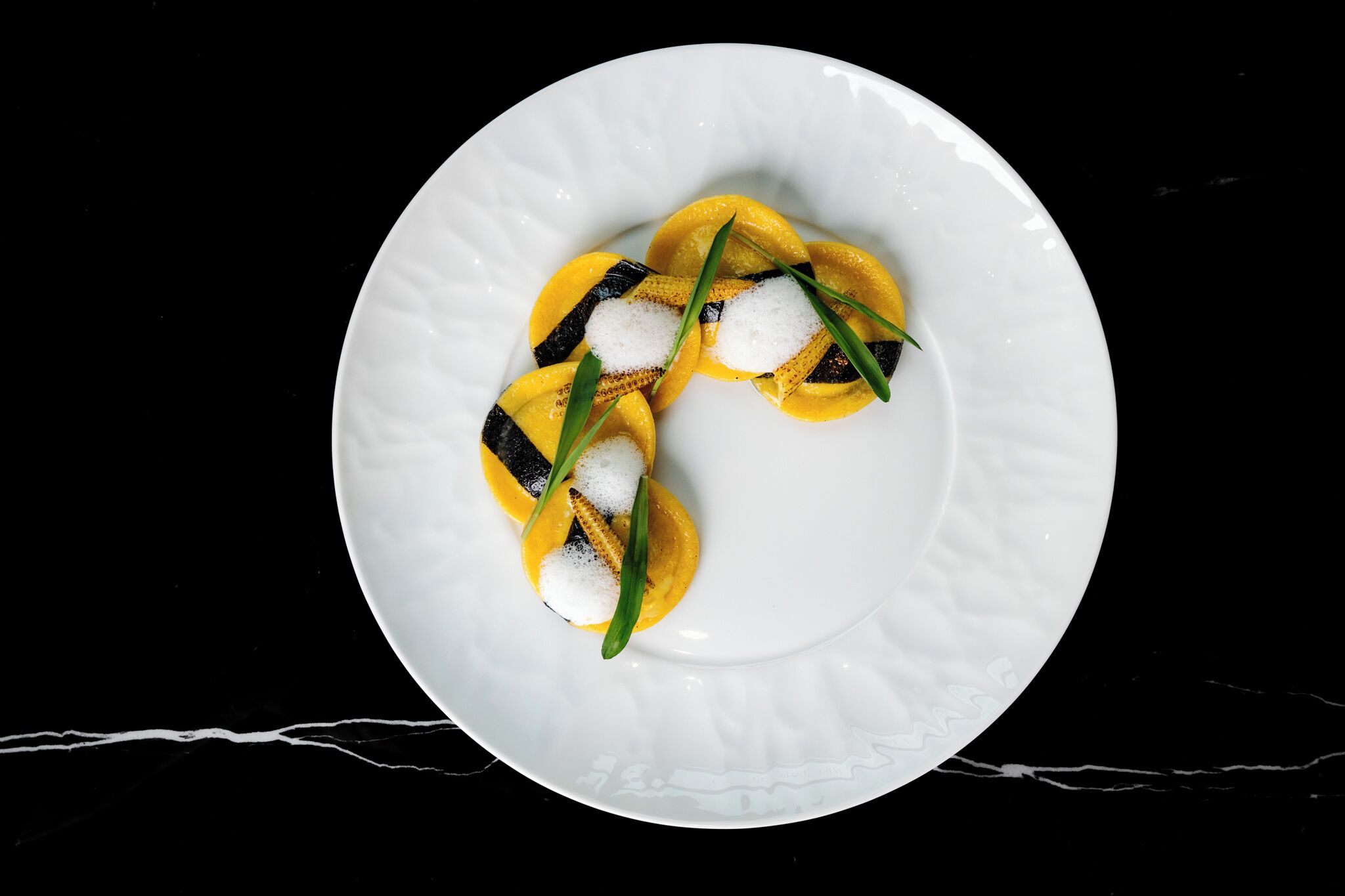 A white plate with a dessert consisting of yellow and black items, garnished with green leaves and white sugar.