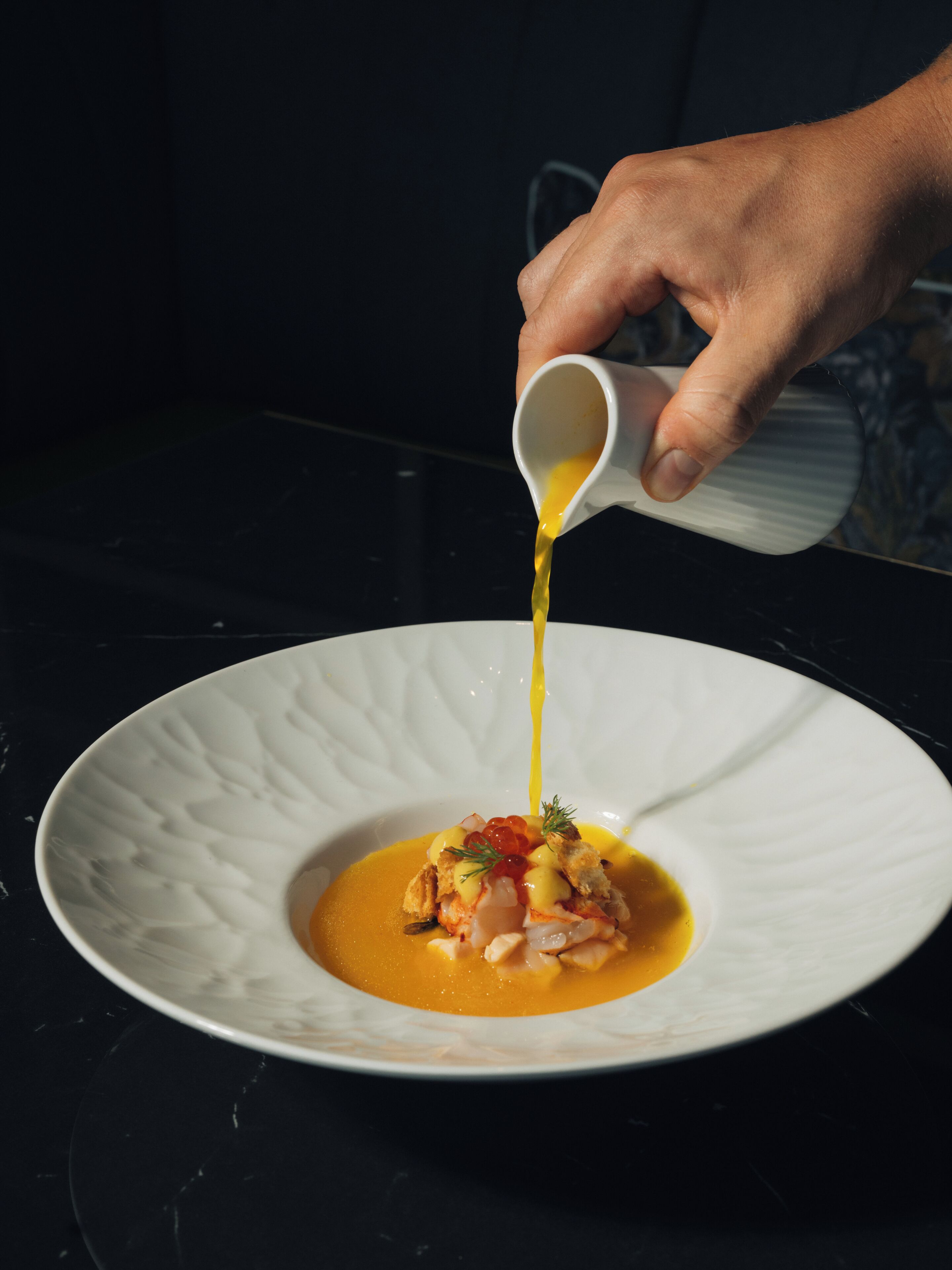 A hand pouring a yellow sauce onto a white plate with a seafood dish.