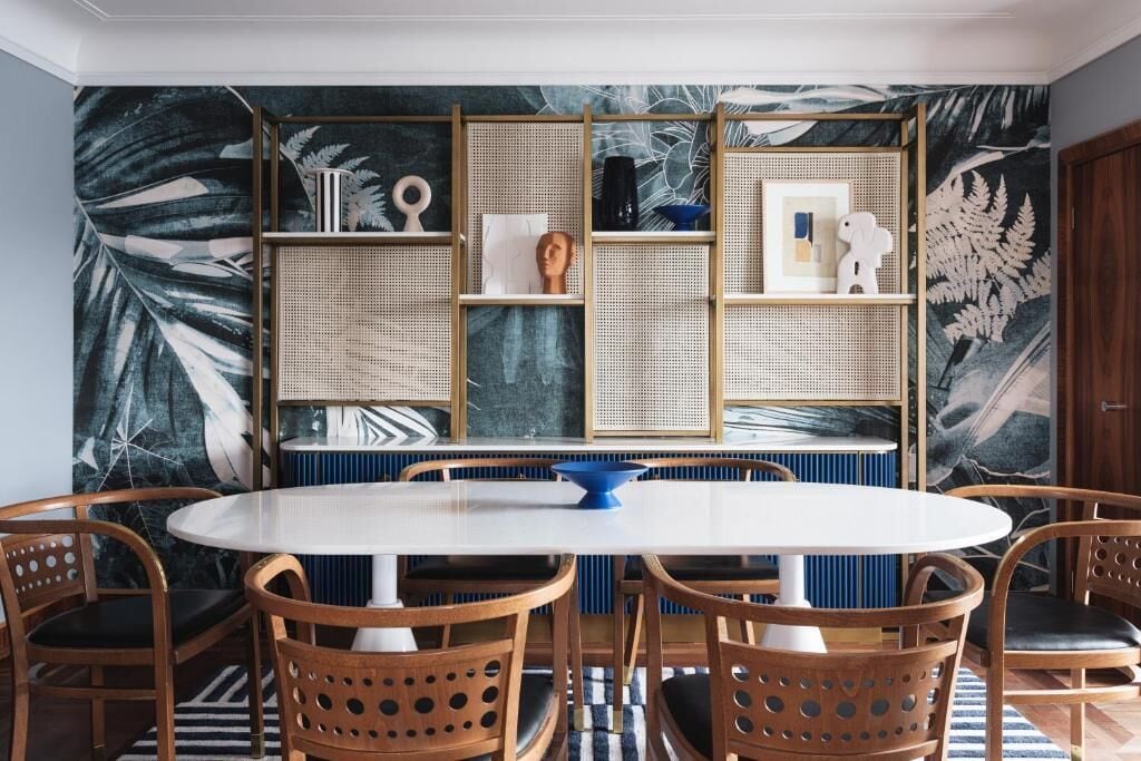 A dining area with a round table and six wooden chairs, surrounded by a wall with a tropical leaf pattern and a woven partition.
