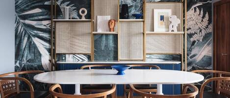 A dining area with a round table and six wooden chairs, surrounded by a wall with a tropical leaf pattern and a woven partition.