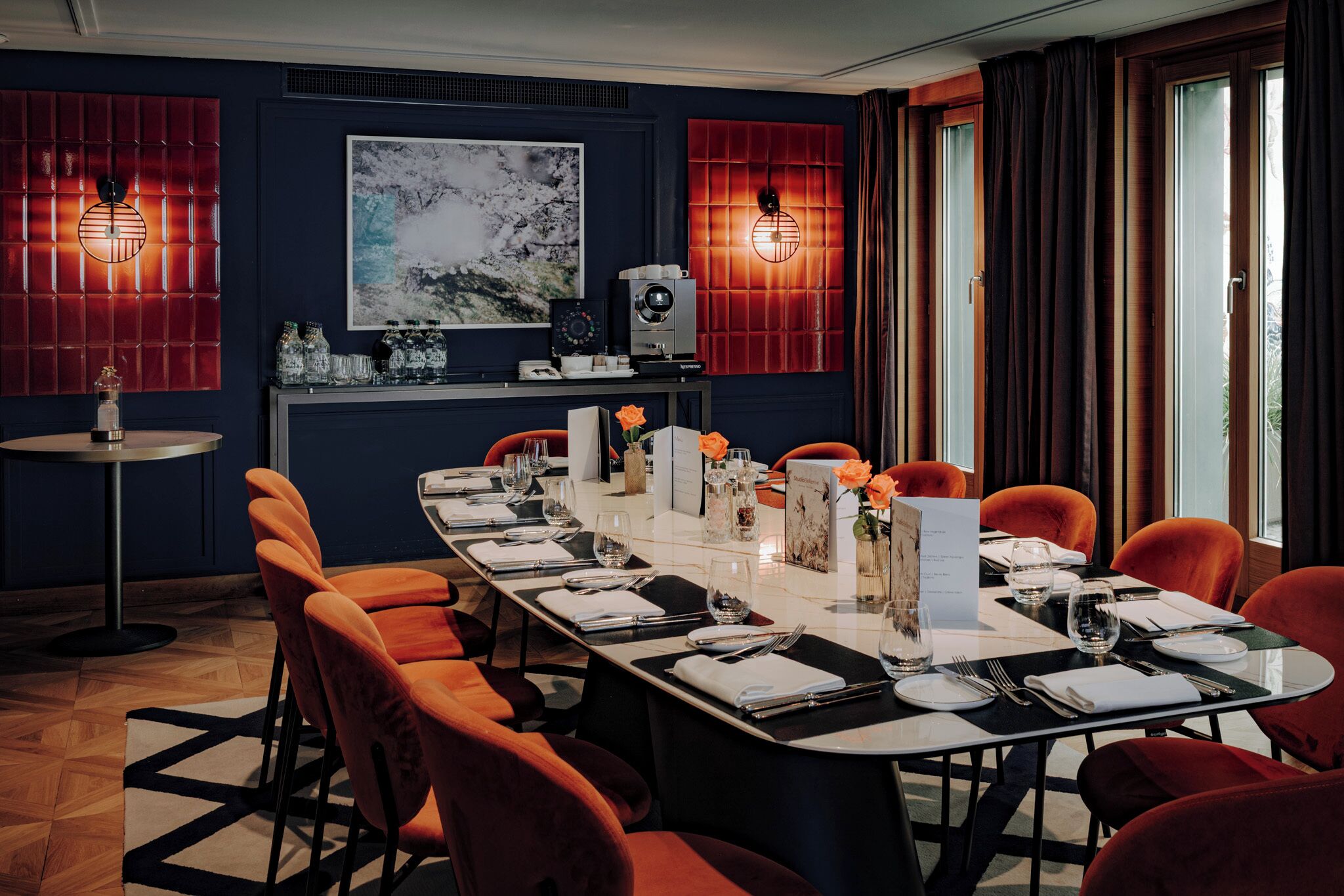 Un espace repas avec une longue table dressée pour un repas, entourée de chaises orange, avec un mur d’accent rouge et une grande peinture abstraite au-dessus de la table.