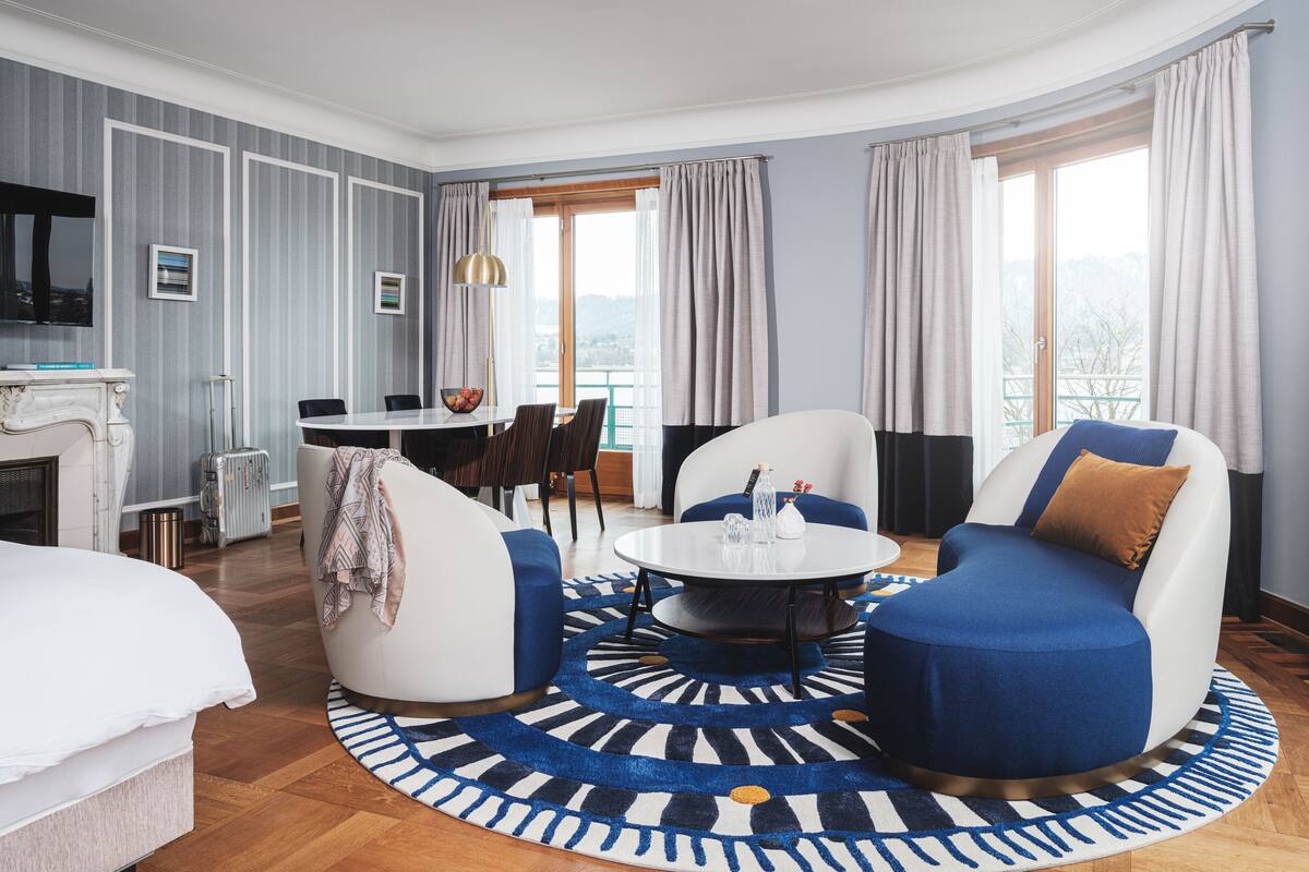 Habitación de hotel moderna con una cama grande, una mesa de centro redonda, dos sofás, un comedor con mesa y sillas, y vistas al exterior a través de amplios ventanales.