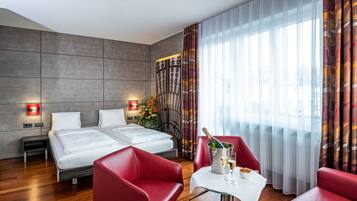 Ein modernes Hotelzimmer mit zwei Betten, einem roten Sessel, einem kleinen Tisch mit einer Champagnerflasche und einer Zimmerpflanze.