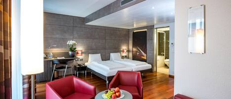 A modern hotel room with a bed, two red chairs, a small table with fruit, a desk, and a nightstand.