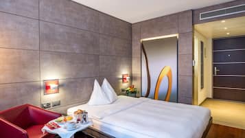 Une chambre d’hôtel moderne avec un grand lit, un fauteuil rouge, une petite table avec des articles pour le petit-déjeuner et un grand miroir.