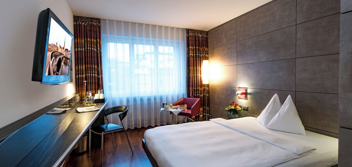 A hotel room with a bed, a desk with a chair, a television mounted on the wall, and a window with curtains.