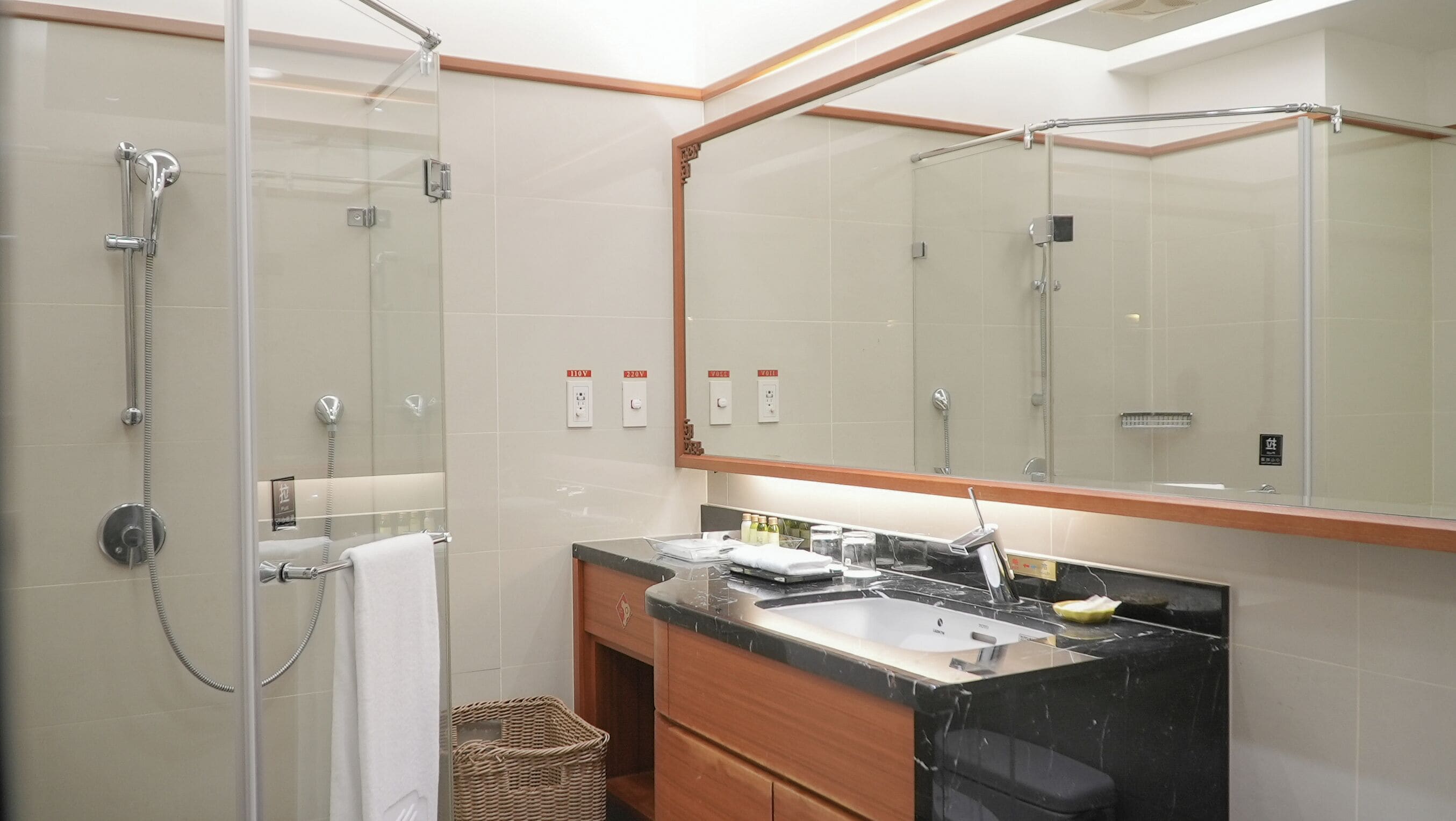 Un baño moderno con ducha acristalada, un espejo grande y tocador con lavamanos y artículos de aseo.