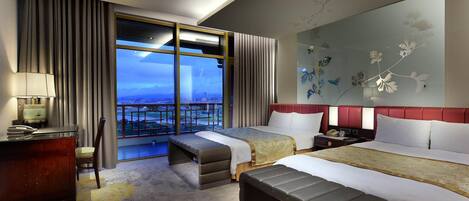 A hotel room with a large bed, a bench, a desk, and a view of a cityscape through a large window.