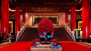 A grand hotel lobby with a red carpet, a staircase, and a large floral arrangement.