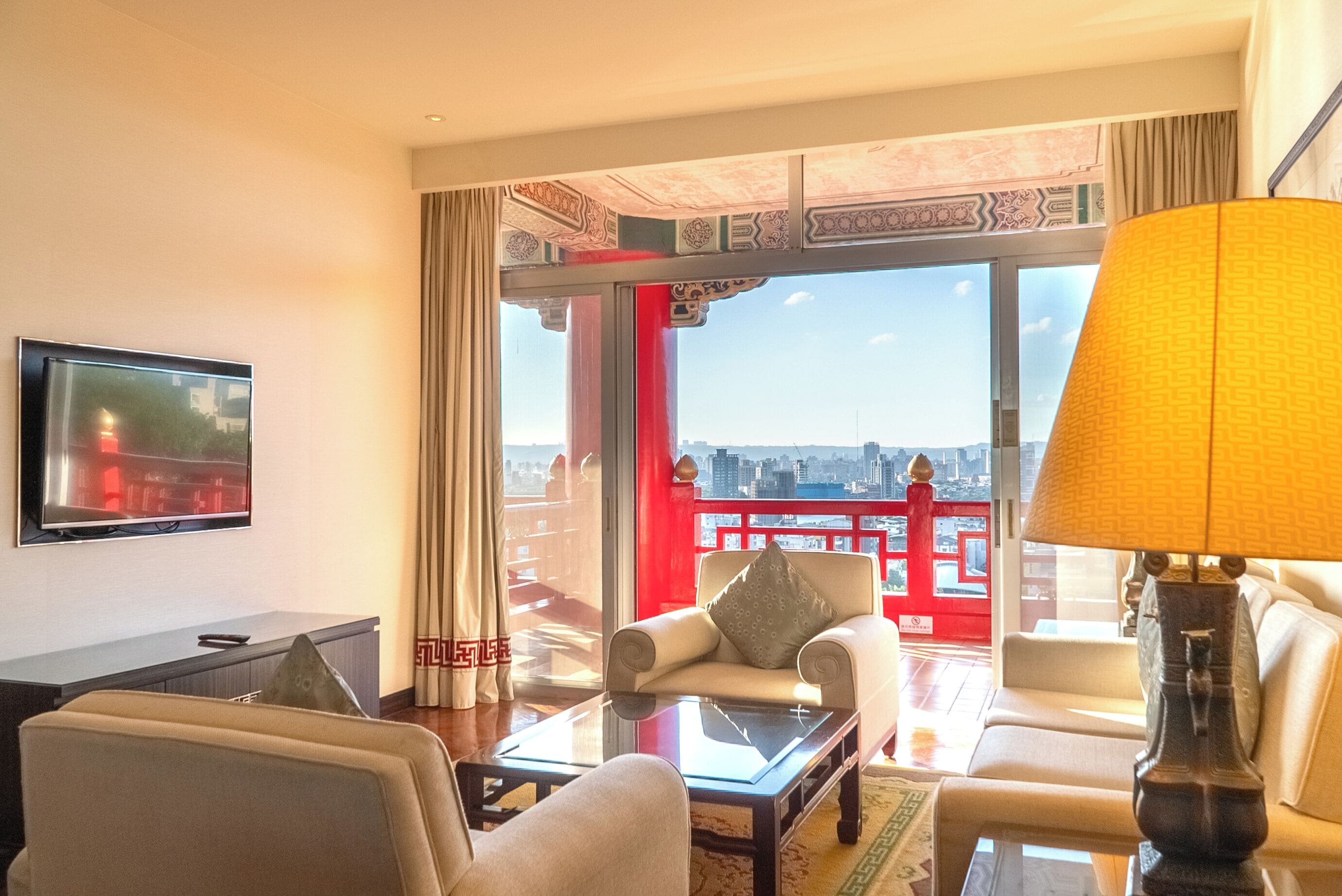 A hotel room with a balcony, a sofa, a chair, a TV, and a lamp.