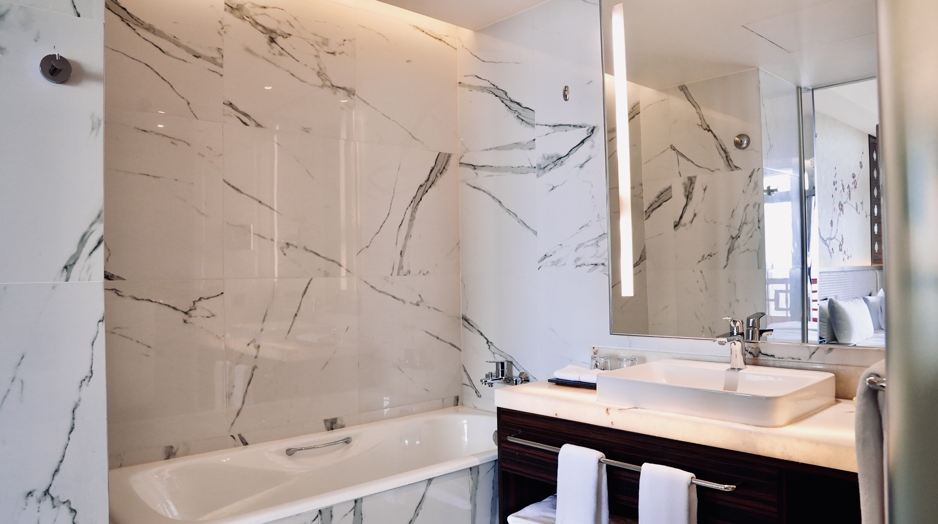 Une salle de bain moderne avec un mur en marbre, une baignoire, un meuble-lavabo avec un miroir et un porte-serviettes.