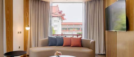 A modern hotel room with a sofa, a coffee table with a cake and flowers, a TV on the wall, and a view of a traditional building outside.