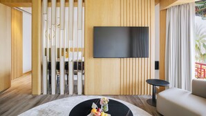 A modern living room with a flat-screen TV mounted on a wooden wall, a round coffee table with fruits and a vase, a sofa, and a view of a balcony with a railing.