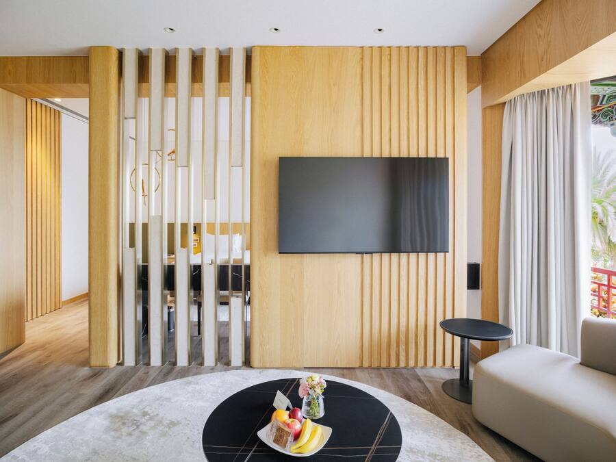 Een moderne woonkamer met een flatscreen televisie aan een houten wand, een ronde salontafel met fruit en een vaas, een bank en een uitzicht op een balkon met een hekwerk.