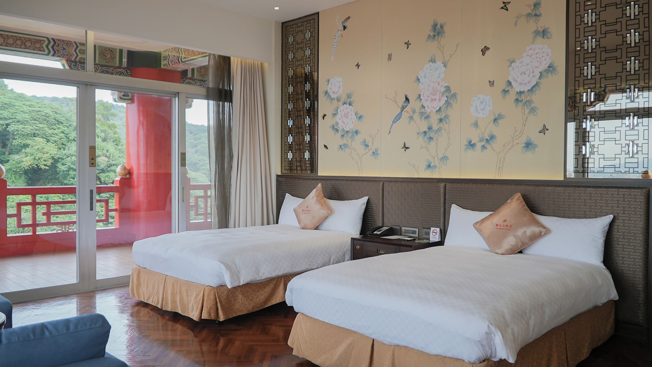 A hotel room with two beds, a large window with a view of greenery, a decorative wall with floral patterns, and a red balcony.