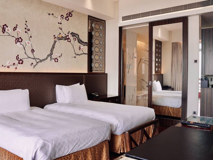 A hotel room with two beds, a large painting of cherry blossoms, a wooden headboard, and a view through an open door.