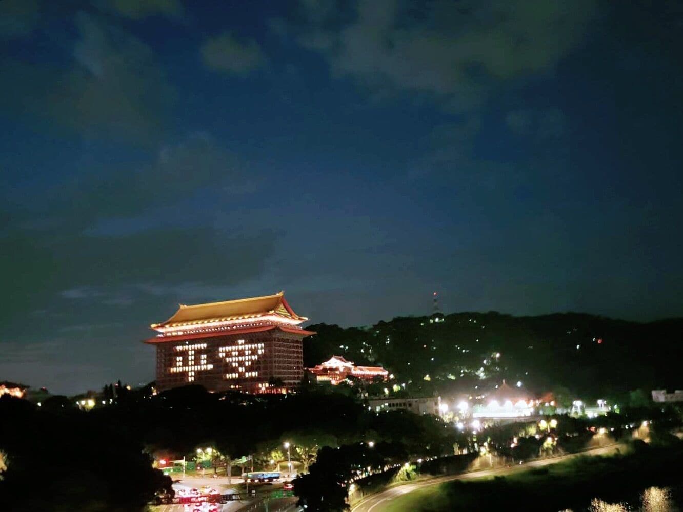A traditional building with a tiled roof and illuminated windows, surrounded by a hilly landscape.