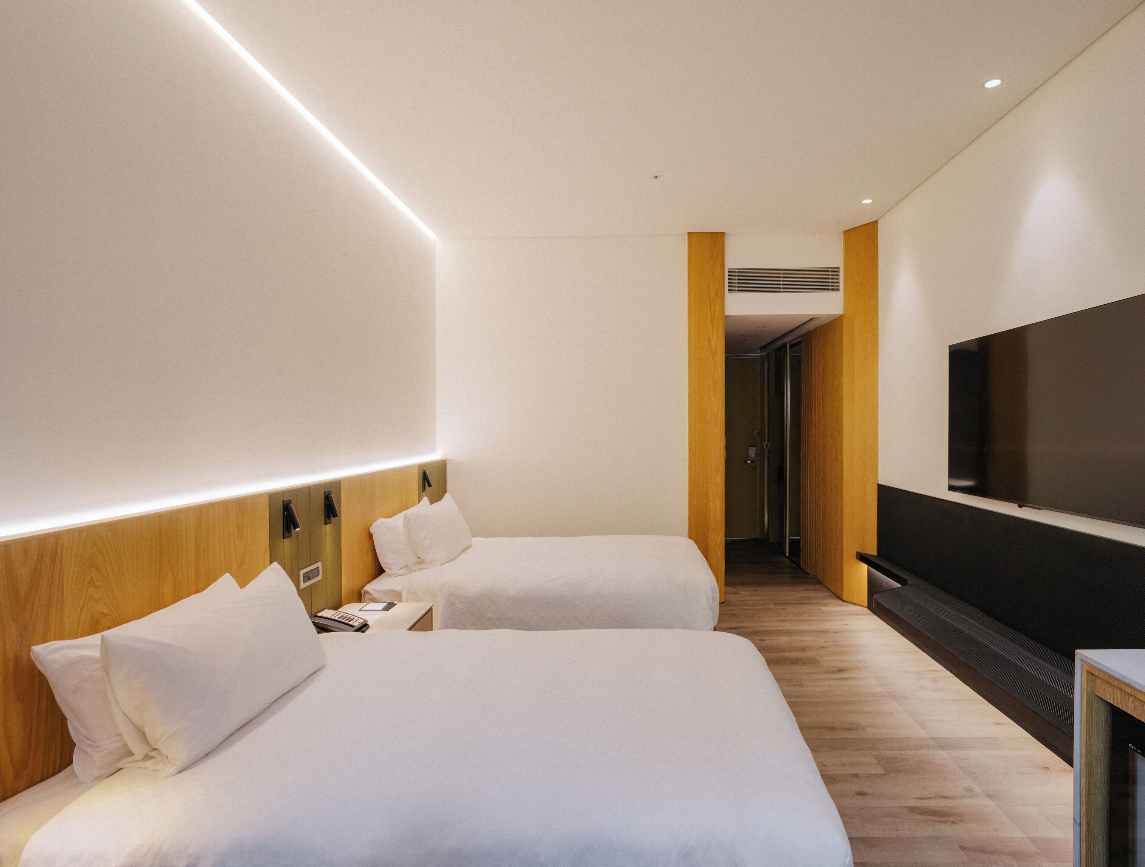 A hotel room with two beds, a flat-screen TV, wooden headboards, and a hallway leading to another room.