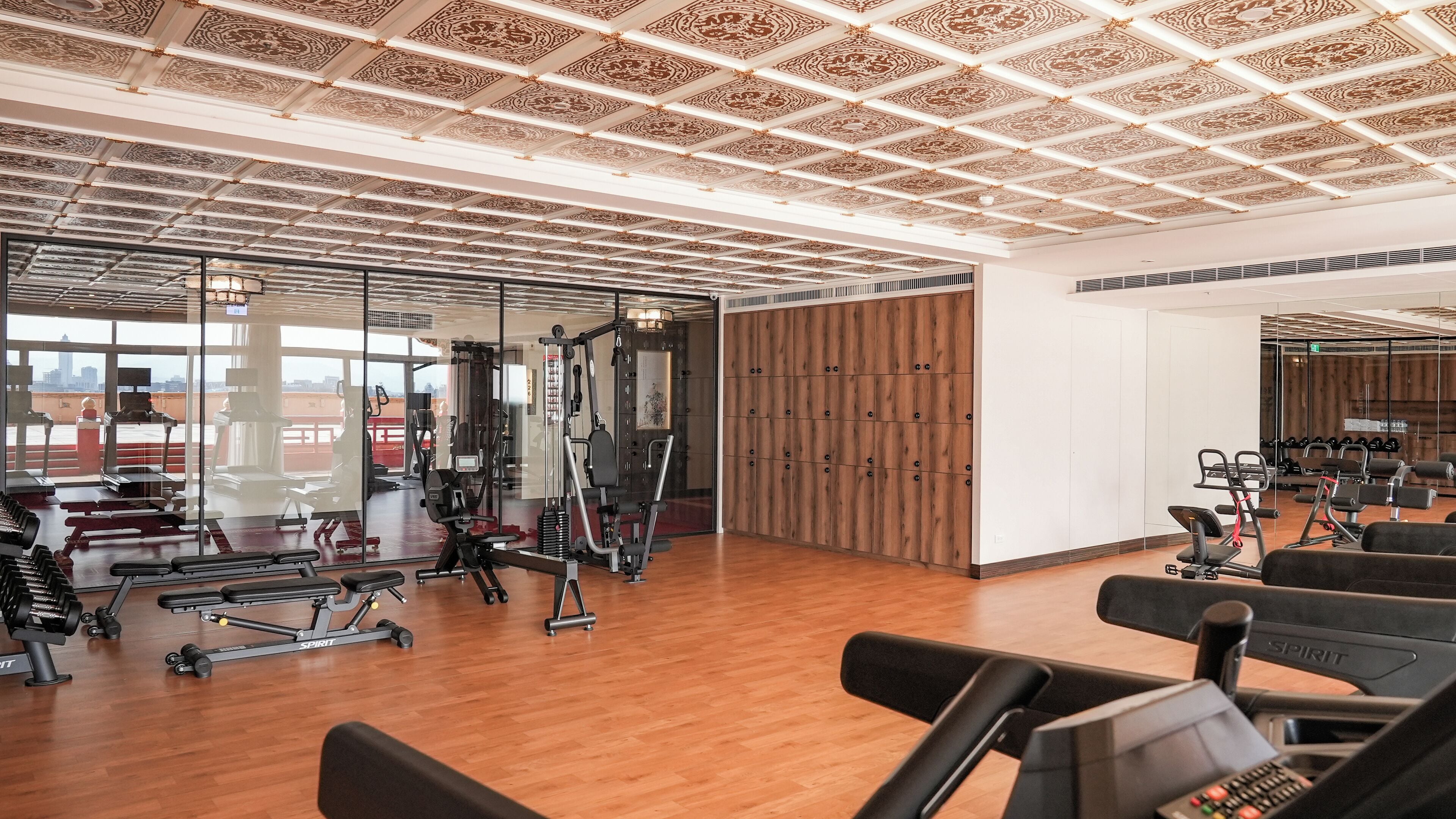 A modern gym with various exercise equipment, large windows, and a wooden floor.